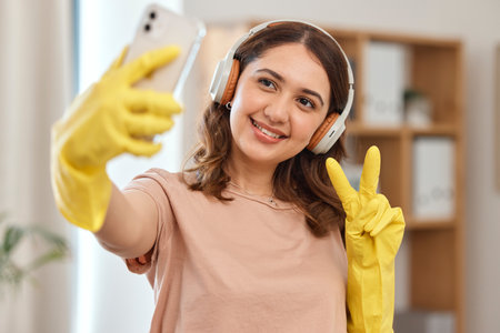 Cleaner woman, selfie and peace sign with smile, headphones and audio streaming for music in home. Happy maid girl, emoji or icon with photography, memory and profile picture on social network appの写真素材