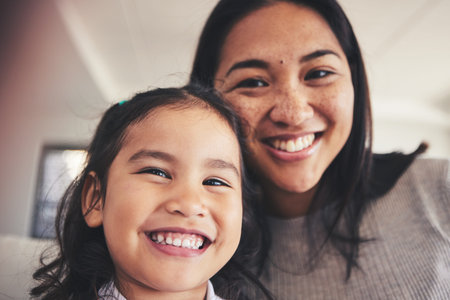 Selfie, smile and portrait of a girl with her mother bonding in the living room of their home. Happy, love and face headshot of Asian child taking a picture with her young mom at their family house.の写真素材