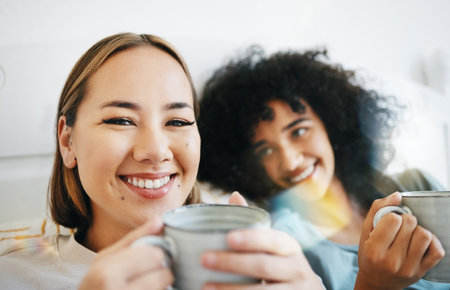 Coffee, smile and portrait of lesbian couple on bed in conversation for bond and relax together. Happy, rest and interracial lgbtq women laughing, talking and drinking latte in bedroom of apartment.の写真素材