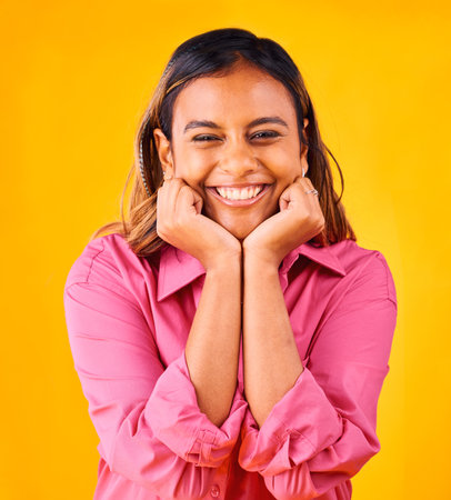 Woman, smile and happy with hands on face, portrait and closeup in studio with yellow background. Indian girl, self love and positive, satisfaction and confidence with hand gesture, style and fashionの写真素材