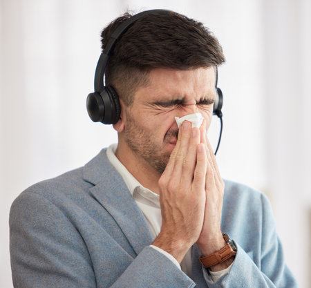 Customer service, sick or man blowing nose in call center office with hay fever sneezing or illness. Cold, contact us or sales agent with toilet paper tissue, allergy virus or flu disease at workの写真素材
