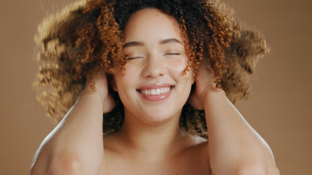 Happy woman, curly hair or natural beauty dermatology for wellness in studio with smile or skin glow. Face, smile or confident biracial female person with skincare cosmetics on a brown backgroundの写真素材