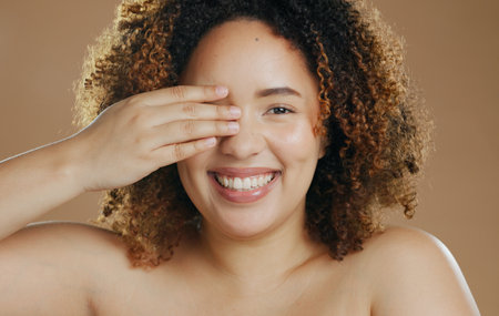 Portrait, funny or happy woman with natural beauty dermatology for wellness in studio with smile. Hand, eye cover or biracial female person laughing with skincare, pride or glow on brown backgroundの写真素材