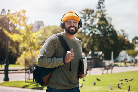 Portrait of happy man on campus with books, headphones and backpack on morning commute for education. Learning, studying and college student in park with podcast, music or streaming online on walk.の写真素材