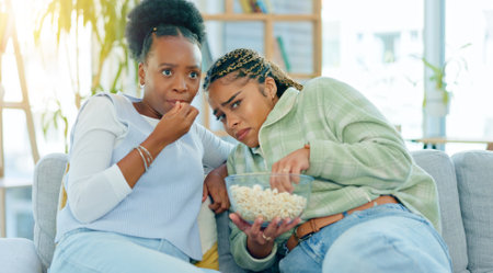 Scary, movie and friends on sofa in living room with popcorn, snack or delicious in house. Black woman, people and together by watching tv, horror or film with expression on face by sitting on couchの写真素材