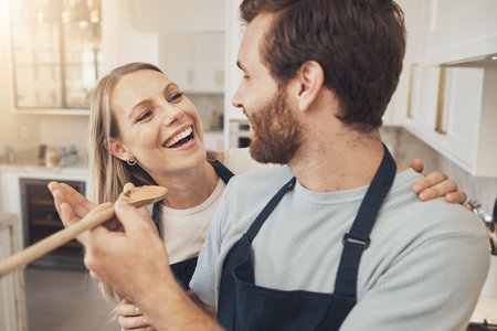 Cooking, taste and happy couple in kitchen bonding while preparing a meal in their home together. Food, spoon and man feeding woman in a house with romance, smile and having fun on weekend or day offの写真素材
