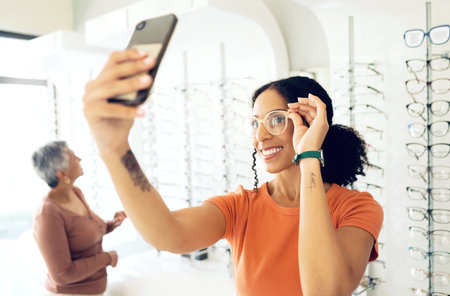 Selfie, optometry and woman trying glasses with prescription lenses and frame in an optical store. Vision, smile and young female person taking a picture on a phone for choosing spectacles in clinic.の写真素材