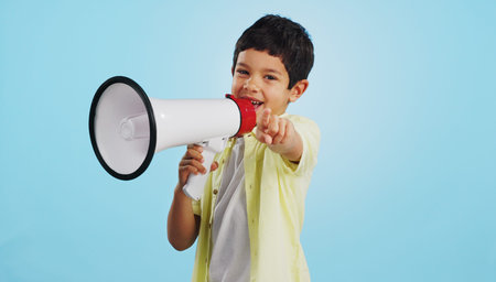Communication, face or child with megaphone for news, opinion or sale announcement on blue background. Happy, pointing or young boy talking on loudspeaker for voice, speaking or attention in studioの写真素材