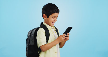 School, phone or kid on social media in studio typing to chat, play mobile games or download app. Blue background, student or happy boy child reading notification for online education or tech searchの写真素材