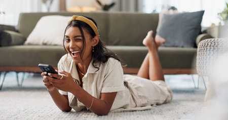 Woman, phone and portrait with wink, relax and lying on floor for flirting, secret and typing in living room of home. Indian, girl and smartphone on ground for chat, texting or technology with emojiの写真素材