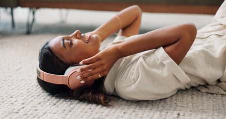 Headphones, relax and young woman in the living room laying on the mat listening to music. Smile, calm and Indian female person streaming podcast, radio or playlist and chilling on the floor at home.の写真素材