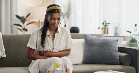 Stomach pain, woman and stress on couch at home with abdomen problem and constipation from virus. Sick person, menstruation and bloated belly ache from colon health issue and cramps in a living roomの写真素材