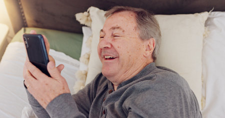Happy, senior man and reading with phone in bed or streaming funny, comedy or meme on social media. Elderly person, smile or relax with cellphone at night in bedroom with communication or online chatの写真素材