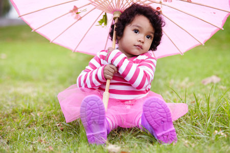 Umbrella, cute and girl child in a garden sitting on the grass on summer weekend. Adorable, playful and young kid, baby or toddler with curly hair playing on the lawn in outdoor field or park.の写真素材