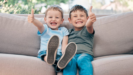 Are we having fun You bet we are. two adorable little boys showing thumbs up on the sofa at home.