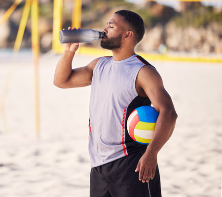 Volleyball, drinking water and fitness of man on beach for training, competition and outdoor game break. Tired, handball and athlete or person with sports bottle on sand in summer or holiday wellnessの写真素材