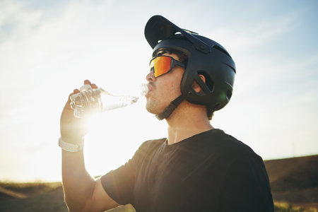 Biker, drinking water and man with helmet and sunglasses outdoor in nature for sports training workout. Sky, thirst or wellness of male person with hydration for off road cycling, travel or adventureの写真素材