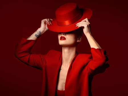 Woman, red aesthetic and hat in studio with makeup art, thinking and mystery with couture, vision and background. Girl, model and high fashion with suit, mindset and facial paint for cosmetic beautyの写真素材