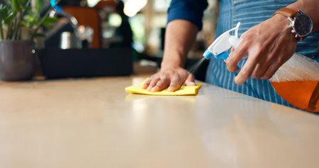 Waiter, hands and cleaning table in restaurant for dust, bacteria and dirt with cloth, spray or detergent. Barista, person or wipe wooden furniture in coffee shop or cafe with chemical liquid closeupの写真素材