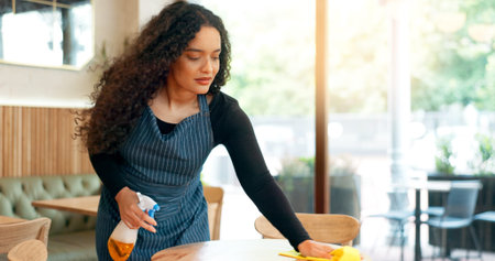 Waitress, woman and cleaning table in restaurant for dust, bacteria and dirt with cloth, spray or...