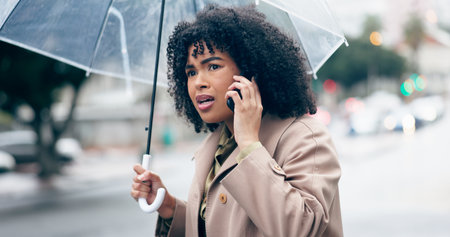 Businesswoman, worry and phone for talking in city for commute to work by walking with umbrella for rain. African person, manager and serious face while late for career, job or meeting with clientの写真素材