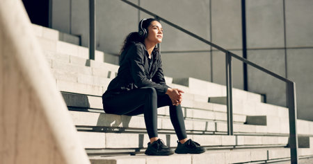 Runner woman, headphones and smile on stairs for music, ideas and relax in city, workout or training. Girl, thinking and happy on steps with audio streaming subscription with peace in summer sunshineの写真素材