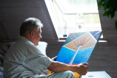 Senior man, artist or holding painting with pride in home by window for creative art or expression. Picture, painter or mature person in room for watercolor drawing or design on canvas for artworkの写真素材