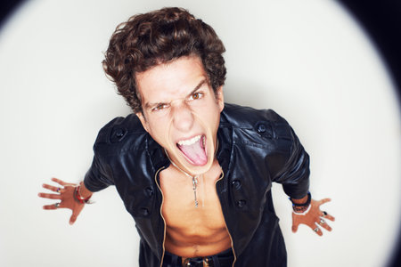 Man, tongue and leather jacket in studio portrait with spotlight, fashion and clothes by white background. Person, top view and rockstar for crazy face, shouting or jewelry for punk aestheticの写真素材
