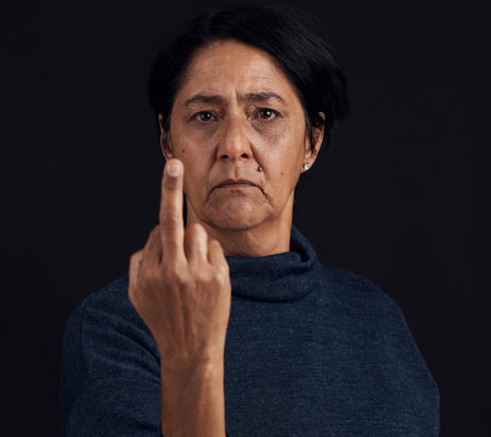 Portrait, anger and senior woman with middle finger, opinion and expression on a black studio background. Face, female person and old model with gesture, rude and frustrated with problem and conflictの写真素材