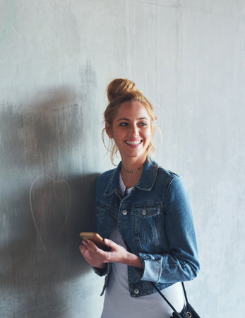 Phone, thinking and smile with a woman student leaning against a gray wall on the campus of her university. Education, idea and mobile with a happy young female pupil standing at college for learningの写真素材