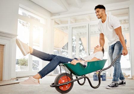 Well always be young at heart. Full length shot of a handsome young man feeling playful while pushing his girlfriend in a wheelbarrow at home.の写真素材