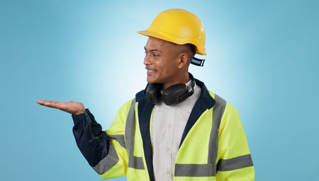 Construction worker, smile and with hand for advertising, promotion or mockup in blue background. Happy, face and builder show safety announcement, presentation or choice with space in studioの写真素材
