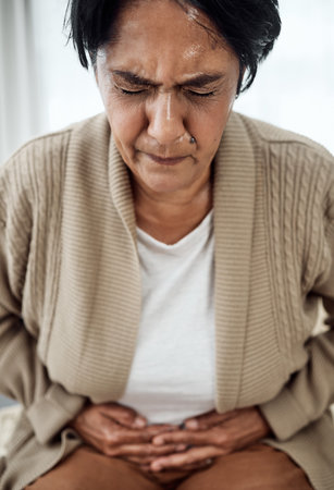Stomach pain, sick or illness with a senior woman in her home, holding her belly in discomfort. Healthcare, medical and emergency with an elderly pensioner feeling uncomfortable after food poisoningの写真素材