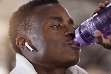 Closeup headshot of an african american athlete drinking water while listening to music through wireless earphones in a gym. Fit, active black man taking break to hydrate during workout and exerciseの写真素材