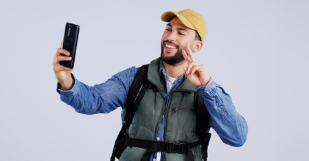 Hiking, smile and man with selfie, peace sign and social media with fun on white studio background. Person, model and hiker with a smartphone, profile picture and adventure with blog, journey or postの写真素材