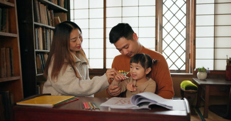 Man, woman and teaching child or books in Japanese for education, learning or development. Family, daughter and pencils at desk in Tokyo for creative art drawing or bonding, kindergarten or writingの写真素材