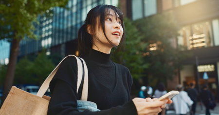 Japanese woman, phone and direction in city with smile, walking or thinking with map by buildings. Person, pedestrian and smartphone with location, schedule or information for address on metro streetの写真素材