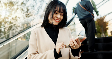Smartphone, happy and Japanese woman on escalator in city, social media app or reading email notification on internet technology in Tokyo. Phone, mobile and person smile outdoor to travel or commuteの写真素材