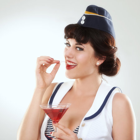 Portrait, stewardess and happy woman with alcohol to drink in studio isolated on white background. Face, martini cocktail glass and air hostess eating cherry, travel or vintage pin up girl on journeyの写真素材