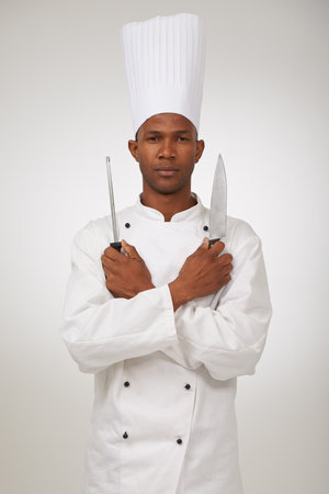 Portrait, african chef and knives in studio in hospitality career and confident cook in food industry. Black man, face and serious in skill in kitchen utensils and uniform by hat by white backgroundの写真素材