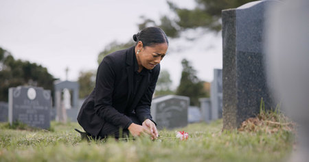 Sad woman, graveyard and crying with rose by tombstone in mourning, loss or grief at funeral or cemetery. Female person with flower in depression, death or goodbye at memorial or burial serviceの写真素材