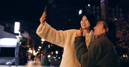Japanese, women and selfie outdoor at night with hand gesture, peace sign and happy in city street. Friends, people and phone with smile for travel, adventure and social media update in urban townの写真素材