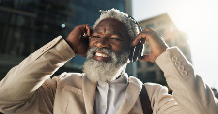 Happy, dancing and senior businessman with headphones in the city walking and listening to music. Smile, happy and elderly African male person streaming playlist, song or radio commuting in town.の写真素材
