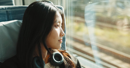 Japanese woman, window and train with headphones, travel and public transportation on metro bullet. Young person, thinking and relax on fast vehicle for weekend trip and wellness by audio technologyの写真素材