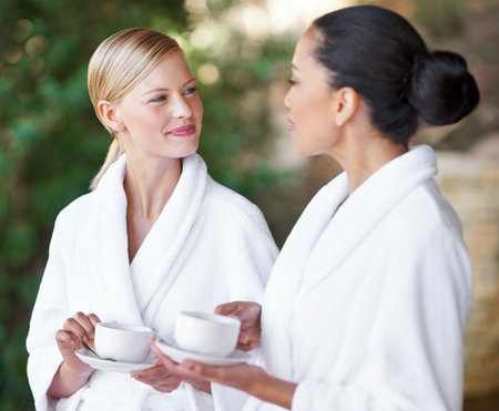Spa, friends or women drinking coffee in nature together for health, wellness or skincare treatment. Girls, tea cup or salon for beauty, luxury body therapy in bathrobe or diverse people talk outdoorの写真素材