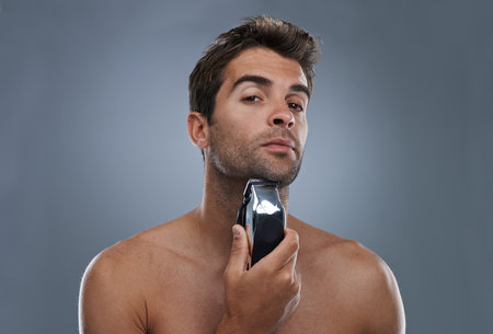 Man, shaving and electric razor in studio portrait for grooming, skincare and wellness by grey background. Person, model and beard with hygiene, facial hair removal and product for cosmetic changeの写真素材
