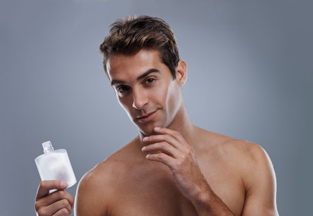 Studio, portrait and man with aftershave for skincare, grooming and facial treatment or cologne. Model, face and happy with bottle for cosmetics product, fragrance and male beauty by gray backgroundの写真素材