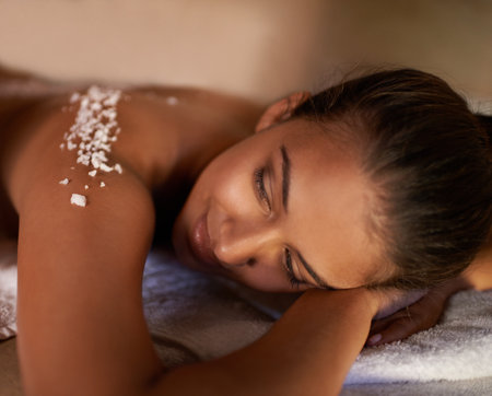 Face, massage and salt with woman at spa on bed or table for luxury pamper treatment or exfoliation. Beauty, peace or skincare and young customer at resort or salon to relax with holistic wellnessの写真素材