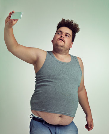 Selfie, fashion and plus size man in studio posing with casual, trendy and cool outfit with stomach. Style, serious and crazy male person with photography picture isolated by gray background.の写真素材
