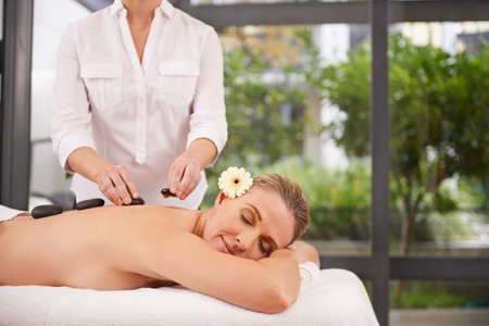 Woman, hands and hot stone massage at spa with masseuse, flower for natural treatment and wellness with zen. Calm, peace and luxury bodycare with warm rocks for detox, self care and holistic healingの写真素材